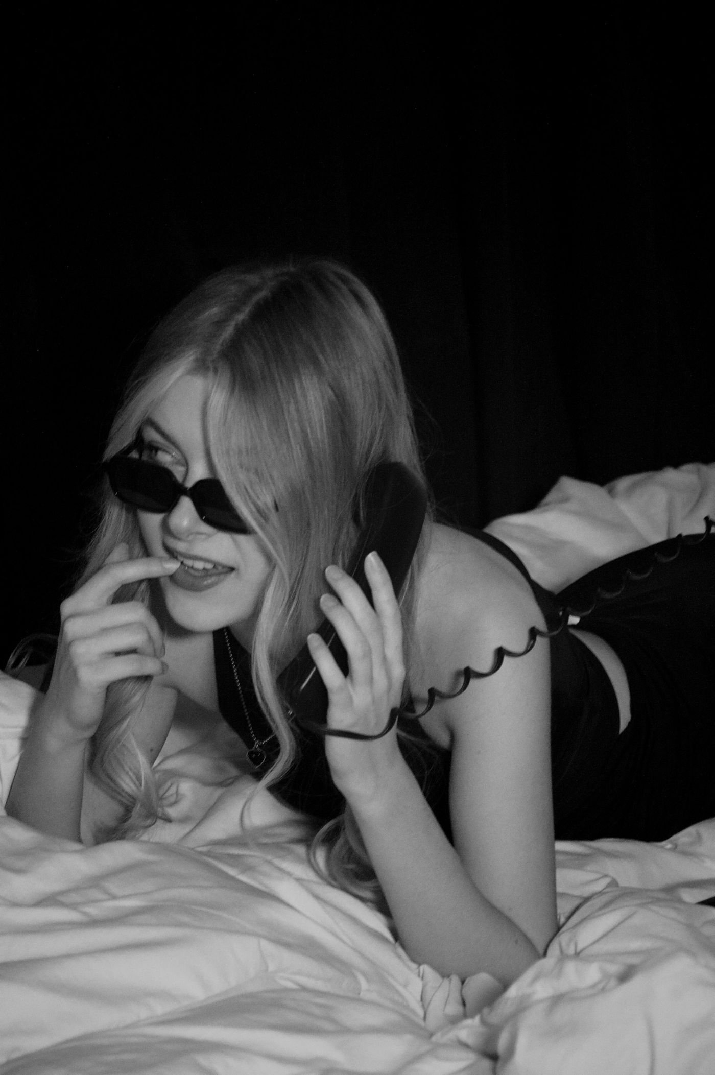 A woman in sunglasses lies on a bed and starts a phone sex call on a vintage phone.